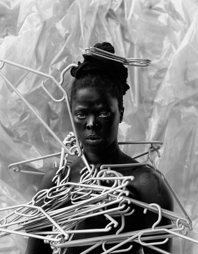 A highly contrasted black and white photograph of Zanele Muholi features the artist gazing out at the viewer, with a mass of silvery coat hangers assembled across their chest and neck.