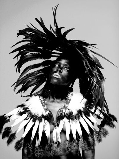 A highly contrasted portrait of Zanele Muholi captures the artist with a feather headdress against a white background, looking up towards the ceiling.