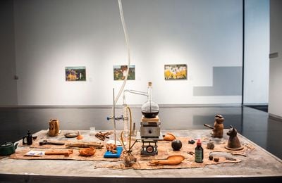 Candice Lin, La Charada China (Tobacco Version) (2019). Cement with casein paint, welded steel table frame, tobacco, ceramics, distillation system (distilling a tincture of tobacco, sugar, tea, and poppy), poppy pod putty, sugarcane, white sugar, cacao, sage, ackee, oak gall, Anadenanthera, dong quai, California clay, Dominican Republic clay, metal parts, bucket, pumps, tubing, dried indigo, glass slides, bottles, drawings, tile, rubber, wood. Exhibition view: The Agnotology of Tigers, LSUMOA, Louisiana (20 October 2021–20 March 2022).