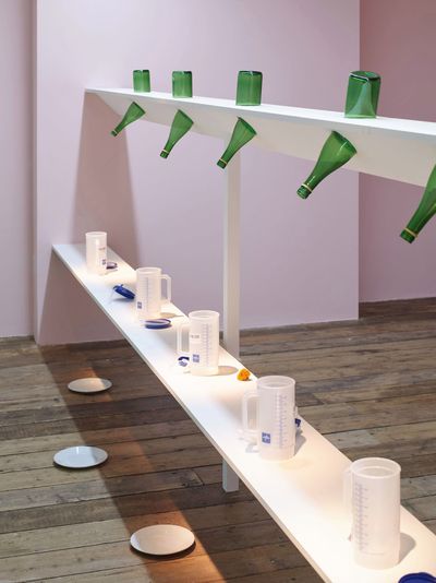 Pope.L, Shelves With Buckfast Bottles (2023). Hospital Carafes And Saucers (Veronica Version), painted wooden shelves, Buckfast bottles, Buckfast wine, isopropyl alcohol, hospital carafes, saucers, and light. Exhibition view: Hospital, South London Gallery, London (21 November 2023–11 February 2024).