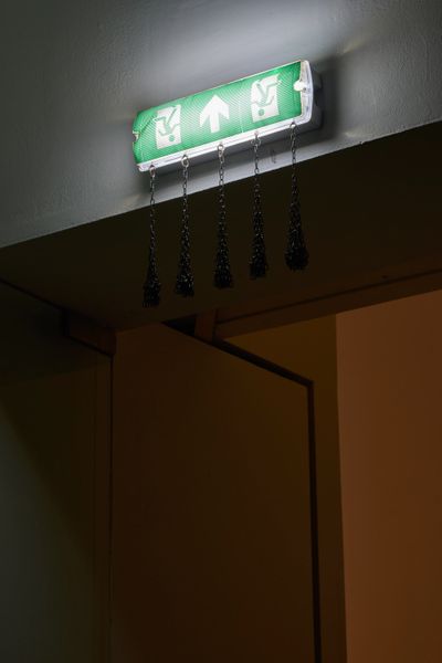 Abbas Zahedi, Exit Sign (2021). Customised exit sign, steel chains, and eye bolts. 40 x 40 x 10 cm. Exhibition view: Horror in the Modernist Block, Ikon Gallery, Birmingham (25 November 2022–1 May 2023).