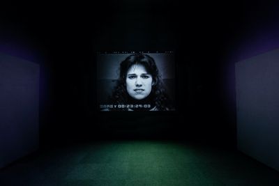 A darkened room shoes a video installation, from which the image of a woman's face is rendered in black and white.