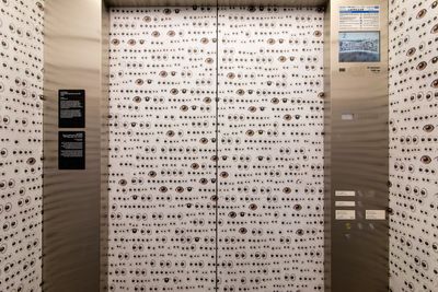 A closed elevator is covered in white wallpaper covered in eyes, as are the walls around the elevator.