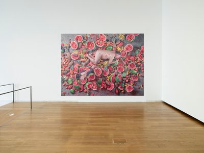 A white wall features a large self-portrait of Ai Weiwei, nude and supine, lying in a foetal position on a bed of battered tropical fruit.