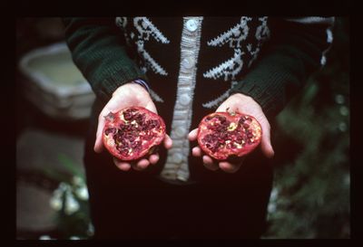 A photograph shows a woman in a black and white sweater, framed from the waist down, holding a split pomegranate, each half shown in either hand.
