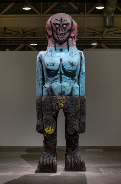 A totemic sculpture by Huma Bhabha that features a many-faced head  painted red, a blue torso, and black legs.