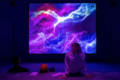 Woman sitting in front of video installation screen projecting waves of electricity static. 