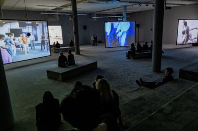 Viewers seated across exhibition space looking at video installation screens. 