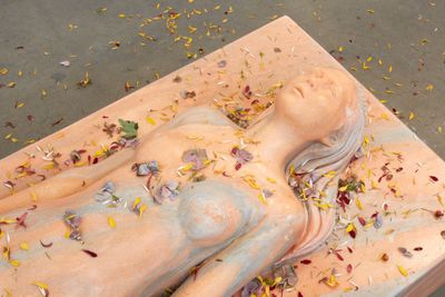 Nude woman covered in petals on the ground made from pink waxed marble and plant matter. 