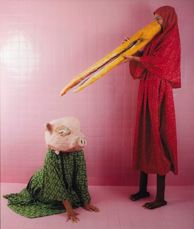Two figures are staged in a pink-tiled room, one sitting on the floor to the left with a papier-mâché pig head, and the other to the right holding a long bird's beak to their face and with feet under her red dress that resemble those of a bird.