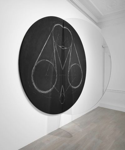 A large circular black canvas with chalk drawn circular motifs by Marco Tirelli is divided in half by a mirror. The sculptural piece hangs in the gallery space.