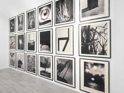Three rows of black and white photographs, with 6 images in each row, hang along a wall in a white gallery space. Each image shows imagery including a tornado, shadows on the ground, and ribbons in the air.