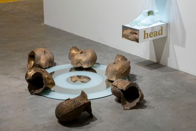Six sculptural heads lie on the floor, surrounding a mirrored plate that sits beneath a pedestal attached to the wall that reads 'head'. Two cast sculptures of feet are placed onto of this pedestal that juts from the wall.