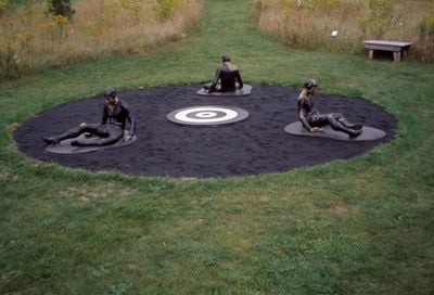 Three sculptures of pilots are seated in a circle in a work by Michael Richards. Each pilot appears to be drenched in tar and stuck to the ground.
