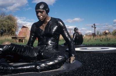An outdoor sculpture by Michael Richards is photographed up close, capturing one of the installation's pilots drenched in tar, seemingly stuck to the ground.