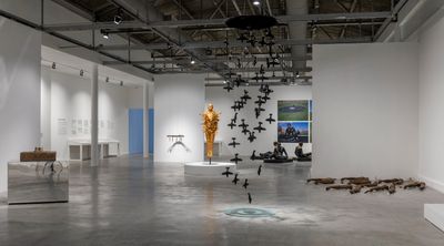 A vast exhibition space features a variety of sculptural works by the artist Michael Richards, including a series of black airplanes cascading from the sky and a golden figure in the distance that is intercepted by airplanes.