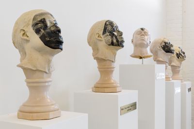 A series of five plaster heads are placed on a plinth each. Each face features black and white newspaper imagery of police in riot gear, except for the central head which features a black and white portrait of activist Rodney King.