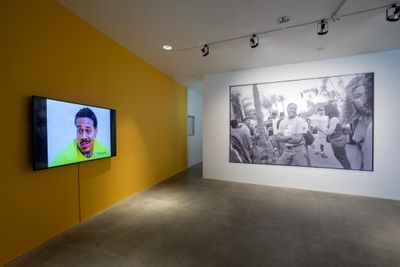 A room in the exhibition space features one yellow wall with a video placed on it, from which a male figure is captured in the midst of speaking. To the right, a white wall features a black and white archival image of the artist Michael Richards.
