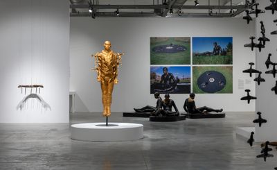 A series of sculptures by artist Michael Richards are placed throughout an exhibition space, including a cascade of sculptural airplanes hanging from the sky, a golden figure with planes intercepting their body like knives, and three pilots seated onto of circular black pedestals.