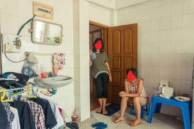 Two women are photographed in a domestic setting, one sitting on a stool while the other stands in a doorway. The two figures, likely women, have their faces covered by red blotches.