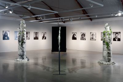 A series of photographs of portraits in black and white hang along the two walls of the gallery space in the background, while stone totem sculptures feature in the foreground, their surfaces featuring some plant life.