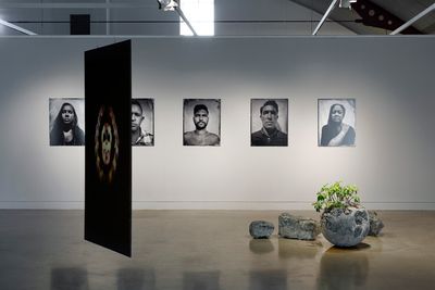 A series of five black and white portraits hang in a line on a  white wall in the gallery space, while a series of five stone sculptures are arranged in a cluster on the floor to the right, and a hanging video projection to the left features hands overlapping into a skull formation.