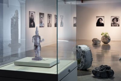 An installation shot of a gallery space shows portrait photographs hanging along the wall, with a silver figure on a pedestal in the foreground and three stone-like sculptures dotted along the floor.