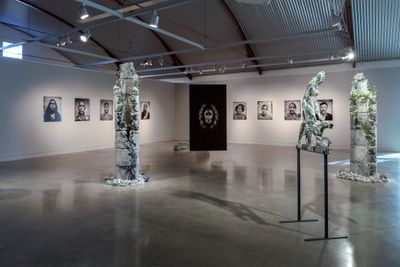 A photograph of at the gallery space features two walls with black and white portraits along them, while totem stone sculptures, plants coming out of their surface, are accompanied by a sculpture of a man and dog in the foreground.