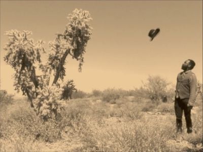 A sepia film still shows Samson Kambalu in a desert landscape, his hat suspended in the air beside him.