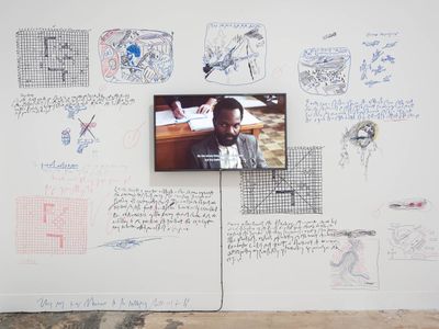 A white wall is filled with inscriptions and scribbles by Samson Kambalu, which surround a video on the wall that pictures the artist sitting in what appears to be a courtroom.