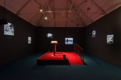 A podium with a red floor is illuminated in the centre of a darkened room. Across the black walls, video projections are arranged.