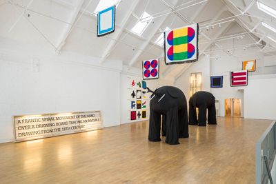 Samson Kambalu's exhibition at Modern Art shows two large black elephants in the middle of a white room with a high ceiling, which also contains abstract, geometric, and colourful flags hung around the space.
