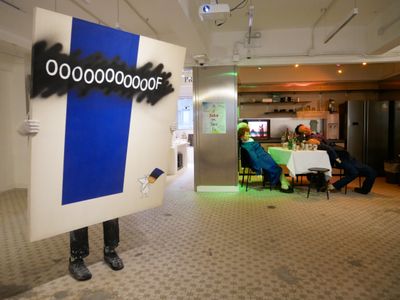 A painting with 'Oooof' written on its surface is held up by white-gloved hands, the figure behind it invisible apart from their feet. In the background of the exhibition space, a table with cans of beer on it is surrounded by dummies slouched on their chairs.