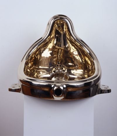 Marcel Duchamp's iconic 'Fountain' sculpture of a urinal is recreated by Sherrie Levine and sits atop a pedestal, its golden surface shining.