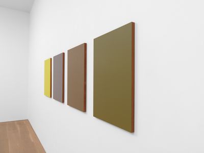 Four panels in shades of khaki, purple, and yellow are photographed at a slant, lining the wall of a white gallery.