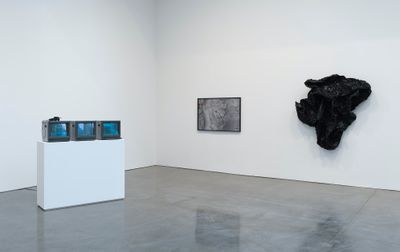 A set of three television sets on a white pedestal in a white gallery space sit to the left of the frame, while two works hang on the wall to the right: one of which resembles a large segment of black rock, while the other looks like a glowing rock surface encased in glass.