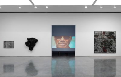A wall in a gallery space features an opening that looks into a darkened room, where a video projection is showing. The video shows the face of a male figure wearing sunglasses. Back in the gallery space, the walls either side of the opening feature three artworks, including two geological wall sculptures to the left, and a painting by Rick Lowe to the right.
