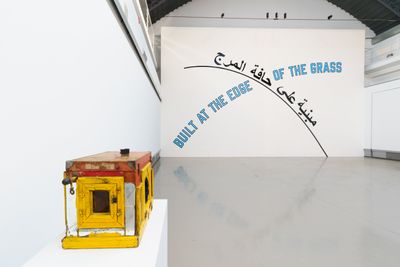 Sound piece and text on wall reading 'Build at the edge of the grass' intersecting with Arabic text.