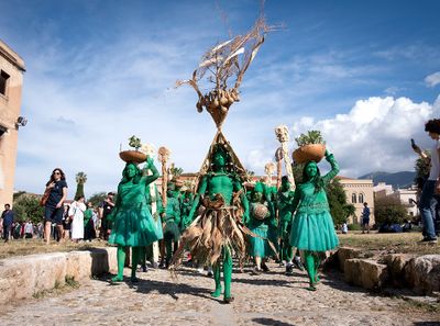 Performance showing procession of bodies painted green parading down dirt road wearing large headpiece.  