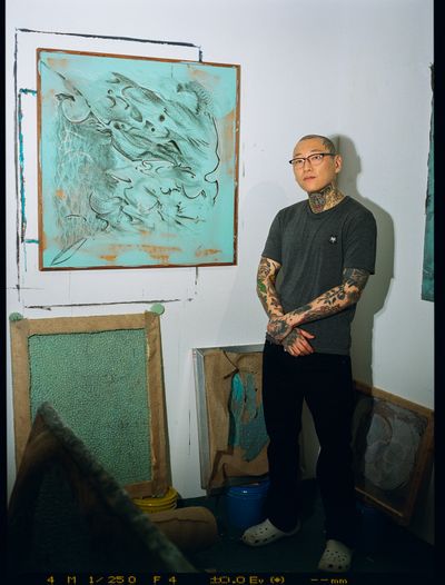 Yissho Oh posing in front of Untitled (2022). Acrylic, tattoo ink, oil stick on silicone on framed wood panel. 33 x 33 x 11⁄4 inches.