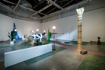 Nabuqi, Do real things happen in moments of rationality?  (2018). Electronic controller, spray-painted FRP cow model, flat car, battery, stainless steel track, outdoor spherical lamp, artificial plant, foam stone, PVC column, inkjet cloth curtain, mirror. 540 x 1600 x 1400 cm. Exhibition view: Nabuqi, Do real things happen in moments of rationality?, ShanghART M50, Shanghi (27 October–9 December 2018). Courtesy ShanghART.
