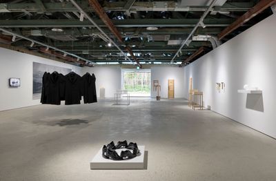 An installation of artworks in the gallery space. Prominent artworks include a small installation of shoes arranged as a circle in the foreground of the exhibition space, while a circle of black coats hangs from the ceiling in the background. 