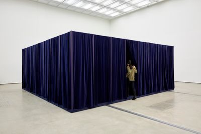 An installation by Ahn Kyuchul in the gallery space features a rectangular structure covered in blue velvet in the gallery space. The image shows an audience member entering the velvet curtains.