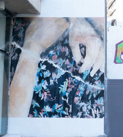 A pair of hands are painted on a mural outdoors, cradling what appears to be a painting of black fabric with blue and red flowers adorning it. The mural is showing outside Galerie Tanit.