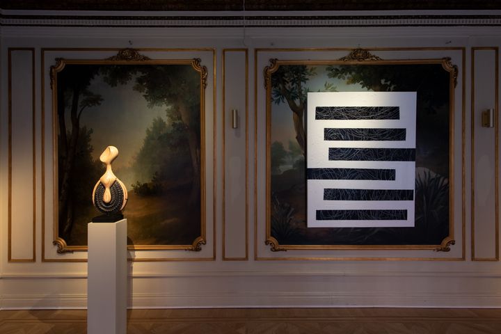 An abstract, curved wooden sculpture is placed on a plinth in the gallery space. To the right of it, there is a black and white abstract painting, made up of stacked bars of class=