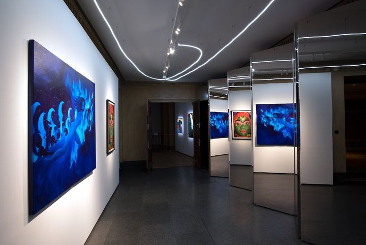 A gallery space shows a blue-hued painting of a female figure running in the moonlight, being chased by hooded class=