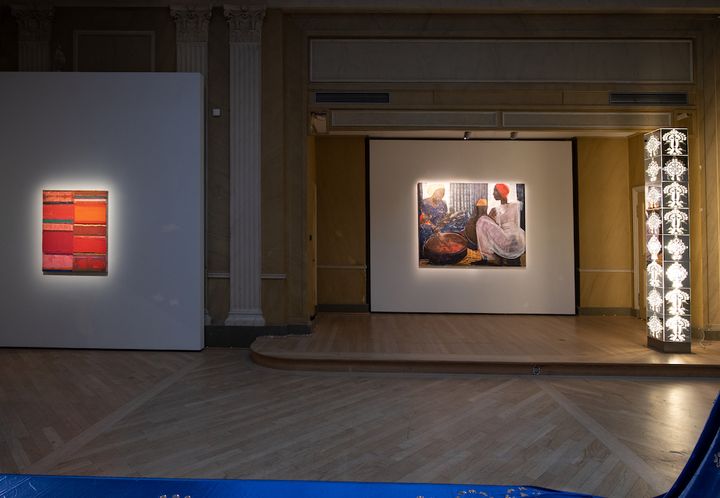A darkened gallery space shows a lit up metallic totem, along with a painting of street vendors and to the far left, an abstract painting of bars of red, pink, yellow, and class=