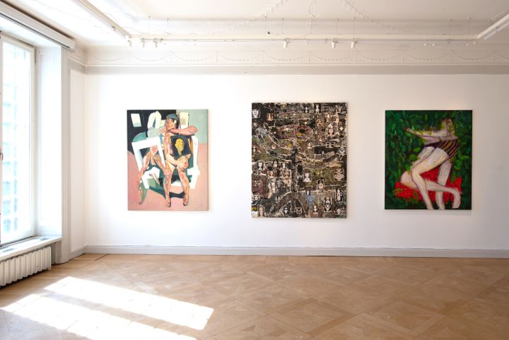A series of three paintings hang on a wall in the gallery space, one depicting two entangled lovers alongside an abstract painting in shades of black and white, and a pink and black-hued painting of a figure sitting on a class=