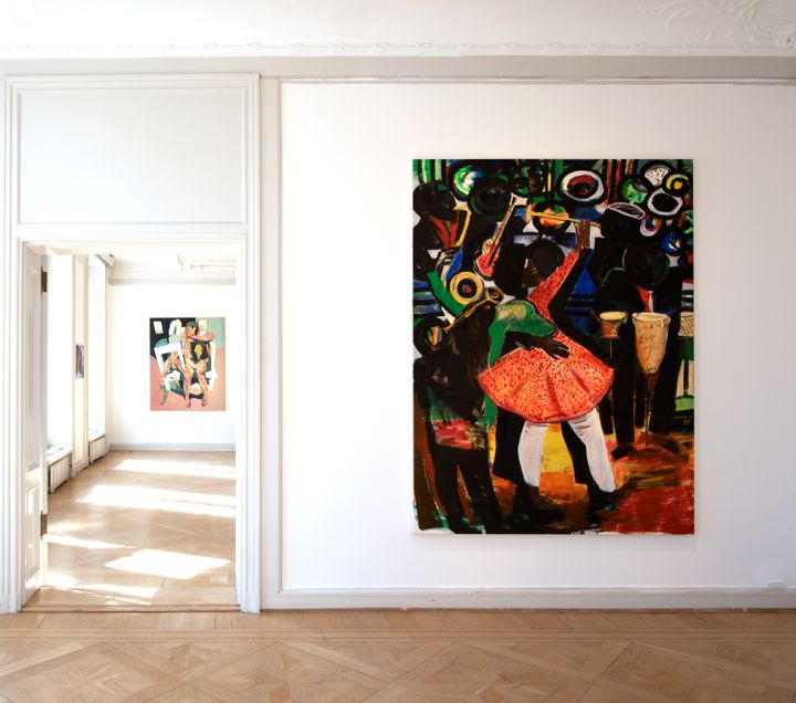 A large painting hanging in a white gallery shoes figures dancing and playing trumpets. In the background, the next-door room is visible, and on it a painting of a fragmentary painting of a figure sitting on a class=