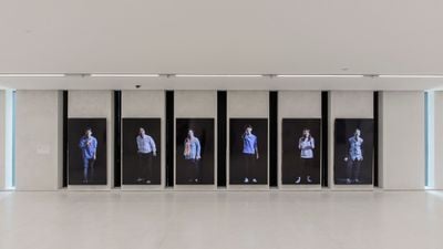 A six-channel video installation by Yang Zhenzhong features six different individuals standing upright on one screen each, each against a black background.